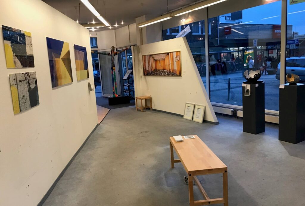 Ausstellungsraum der Pop-up-Galerie mit abstrakten Bildern an der Wand, Skulpturen auf Sockeln und Blick auf die Straße