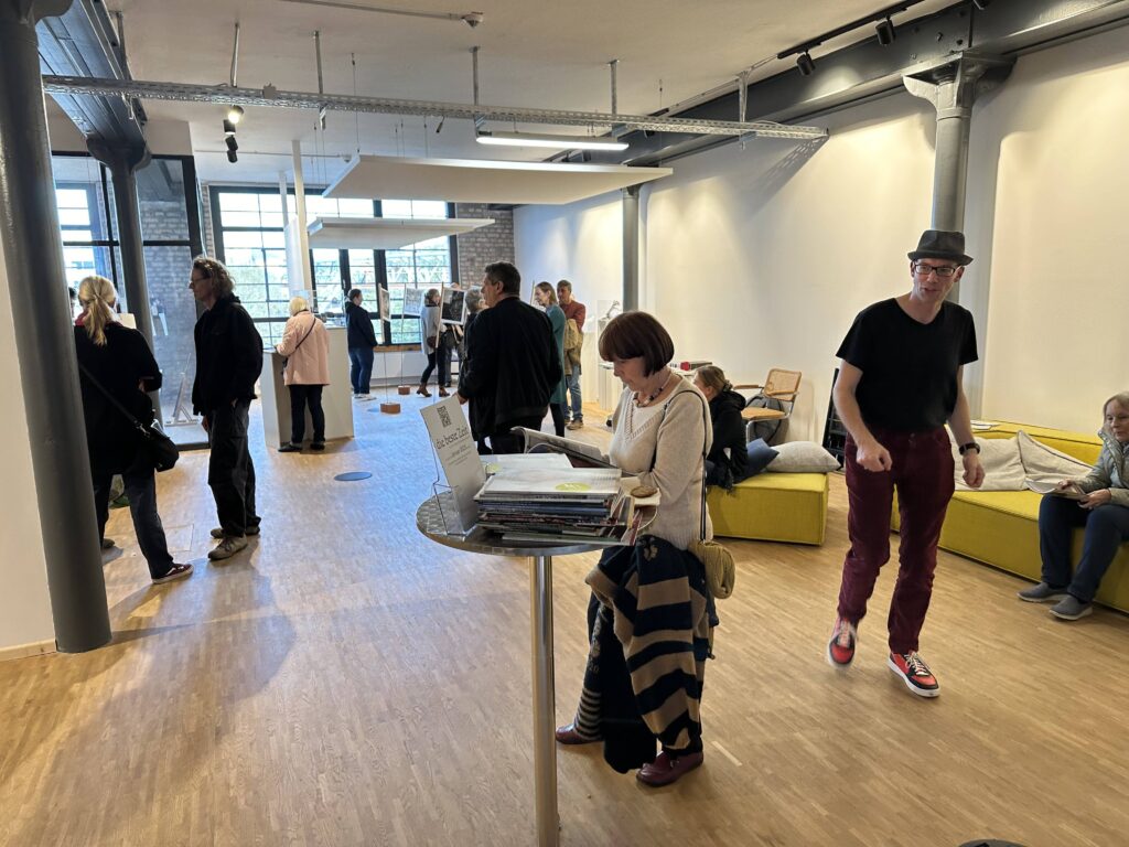 Besucherinnen und Besucher in der Galerie mit Holzboden, gelben Sitzwürfeln und einem Tisch mit Katalogen